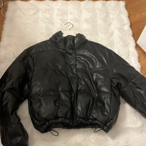 Svart pufferjacka i skinnimitation från shien storlek M - Säljer en svart pufferjacka med glansig skinnlook och hög krage. Jackan har dragsko nertill, tryckknappar framtill och quiltad design. Perfekt för dig som vill ha en trendig och varm jacka med streetkänsla.