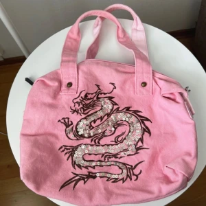 Rosa handväska med paljett-drake - Cool rosa handväska i bomull med dubbelhandtag och dragkedja. Framsidan har en broderad drake dekorerad med glittriga paljetter för extra attityd. Perfekt för dig som vill sticka ut med en unik accessoar.