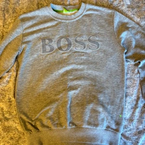 Grå sweatshirt från Hugo Boss - Snygg grå sweatshirt från Hugo Boss med broderad logga framtill. Tröjan har rund halsringning, långa ärmar och ribbade muddar vid ärmslut och nederkant. Perfekt för en clean och stilren look.