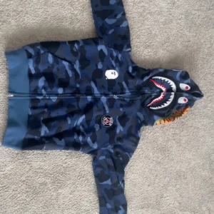Blå camo hoodie från BAPE Shark - Säljer en blå camo hoodie från BAPE med ikonisk shark-hood och dragkedja hela vägen. Hoodien har BAPE-logga på bröstet och detaljer i vitt, rött och orange på huvan. Perfekt statement-plagg för streetwear-stilen.