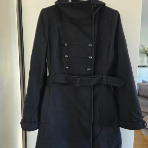 Svart kappa från Stockh LM med bälte - Stilren svart kappa från LM Stockholm  med dubbelknäppning framtill och bälte i midjan. Snygga knapprader som ger en klassisk look. Perfekt för kyliga dagar och passar till många outfits. 