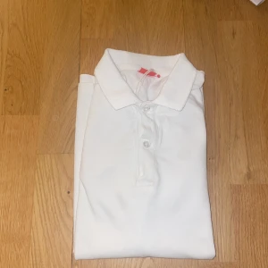 Vit kortärmad piké från RYVLS - Klassisk vit pikétröja från RYVLS med korta ärmar och krage. Tröjan har knappar framtill och är tillverkad i mjuk bomull, perfekt för en clean och fräsch look. Enkel att matcha med jeans eller shorts.