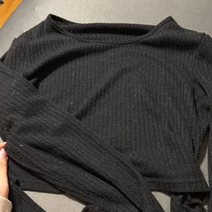 Svart ribbad långärmad topp - En svart långärmad topp med ribbad struktur. Modellen är enkel och tight, perfekt att styla med jeans eller kjol. Toppens material känns mjukt och stretchigt, vilket gör den bekväm att bära hela dagen. Går att knyta vid ryggen! 