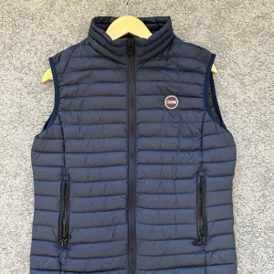 Mörkblå dunväst från Colmar - Snygg mörkblå dunväst från Colmar Originals med quiltad design och hög krage. Västen har två praktiska fickor med dragkedja och ett broderat Colmar-märke på bröstet. Perfekt att slänga över en hoodie för extra värme under höst och vinter.