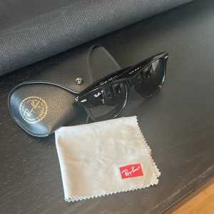Svarta Ray-Ban Wayfarer solglasögon - Klassiska svarta Ray-Ban Wayfarer solglasögon med blank finish och ikonisk fyrkantig form. Kommer med original Ray-Ban-fodral i svart läder och putsduk. Perfekta för dig som vill ha en tidlös och cool look. Pris är ej hugget i sten 