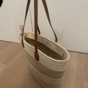 Beige strandväska från Akè  - Snygg strandväska från Akè i beige och vitt. Väskan har långa bruna handtag i läderimitation. Perfekt storlek för att få plats med allt du behöver och stängs med dragkedja upptill.