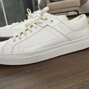 Vita sneakers från Hugo Boss - Snygga vita sneakers från Hugo Boss med stilren design och diskret logga på sidan. Skorna har snörning, platt sula och är tillverkade i skinn. Perfekta för dig som gillar en clean och klassisk look. Små röda detaljer bak ger en extra touch.