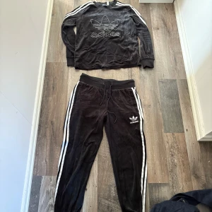 Svarta Adidas mjukisbyxor med ränder - Svarta mjukisbyxor från Adidas med klassiska vita ränder längs sidorna och broderad logga på låret. Byxorna har snörning i midjan och är gjorda i mjuk velour för extra komfort. Perfekta för en avslappnad stil. Även matchende tröja till.