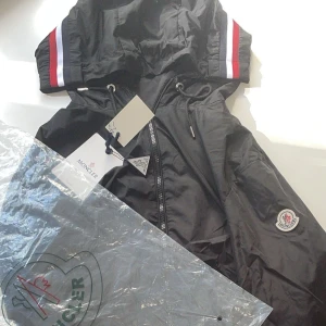 Svart vindjacka från Moncler med huva - Svart vindjacka från Moncler med huva och dragkedja framtill. Jackan har snygga rödvita ränder på huvan och Moncler-logga på ärmen. Justerbar snörning vid midjan och lätt, glansig polyester som ger en sportig vibe. Perfekt för dig som gillar stilrena och exklusiva plagg. Pris kan diskuteras. Passar i M