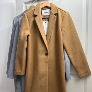 Kamelbrun kappa från ONLY - Säljer en stilren kamelbrun kappa från ONLY med klassisk krage och en knapp framtill. Lång modell med raka ärmar och snygg passform. Perfekt för dig som vill ha en tidlös och trendig look.