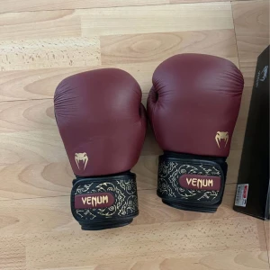 Vinröda boxningshandskar från Venum - Snygga vinröda boxningshandskar från Venum med guldfärgade detaljer och logga. Handledsbanden är svarta med guldmönster och Venum-tryck. Perfekta för dig som vill sticka ut på träningen och köra hårt i ringen. NYPRIS 1199