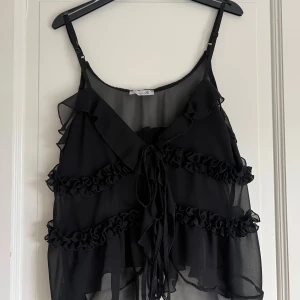 Svart volangig linnetopp med snörning - Säljer en svart, transparent linnetopp från Design by Si med volangdetaljer och snörning framtill. Toppen har tunna, justerbara axelband och är tillverkad i ett lätt, luftigt material som ger en flowy känsla. Perfekt för dig som gillar en edgy och romantisk stil.
