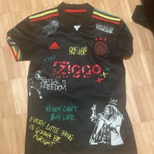 Ajax x Bob Marley svart fotbollströja - Ajax x Bob Marley limited edition fotbollströja i svart med röda, gula och gröna detaljer. Tryck med Ajax-logga, Ziggo, Bob Marley-motiv och inspirerande texter. Korta ärmar med färgglada ränder och broderade detaljer. Materialet är lätt och andas, perfekt för match eller streetwear.
