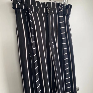 Svarta randiga byxor med knytband - Svarta byxor med vita vertikala ränder och högt papperspåse-midja. Byxorna har ett brett knytband i midjan och raka ben. 