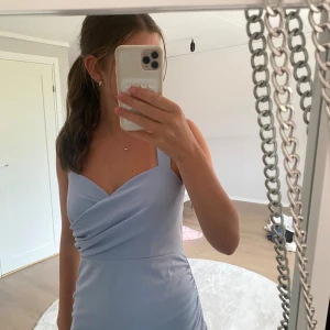 Ljusblå omlottklänning med drapering - Supersöt ljusblå klänning med omlott-detalj och drapering framtill. Klänningen är kort och har breda axelband, vilket ger en stilren och modern look. Perfekt för dig som gillar en feminin och fräsch stil.