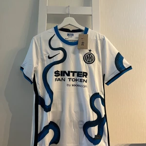 Inter Milan bortatröja Nike S - Inter Milan bortatröja från Nike i vitt med blå och svarta ormliknande detaljer. Tröjan har klubbmärke, sponsortryck och blå ribbade muddar vid ärmslut och krage. Tillverkad i lätt och ventilerande polyester, perfekt för match eller träning.