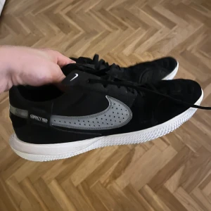 Svarta Nike sneakers med grå swoosh - Snygga svarta Nike sneakers med grå swoosh och vit sula. Skorna har snörning och är tillverkade i mocka och syntet. Swooshen är perforerad och det finns reflekterande detaljer på hälen. Perfekta för dig som gillar stilrena och sportiga skor.