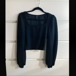 Svart kort långärmad topp  - Svart kort topp med långa ärmar och bred, fyrkantig urringning. Toppens material är mjukt och stretchigt, perfekt för en trendig och enkel look. Passar till både jeans och kjol för en stilren outfit.