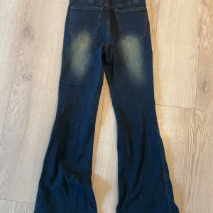 Mörkblå bootcut jeans från Cider - Snygga mörkblå bootcut jeans från Cider med slitningar på låren och baksidan. Jeansen har hög midja, klassiska bakfickor och dekorativa knappar längs utsidan av benen. Tillverkade i stretchig denim för en bekväm passform. Använd endast en gång. 