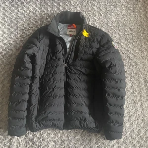 Parajumpers jacka - Svart pufferjacka från Parajumpers med klassisk patch på ärmen och orange detalj vid kragen. Jackan har quiltad design, hög krage och dragkedja framtill. Perfekt för kalla dagar och riktigt snygg till streetwear-stilen.