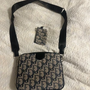 Dior axelväska med monogrammönster - Snygg axelväska från Dior med klassiskt blått monogrammönster på beige botten. Väskan har bred svart axelrem med Dior-logga, silverfärgade dragkedjor och detaljer i skinn. Medföljer matchande liten plånbok/ficka. Perfekt för dig som gillar lyxig streetstyle.