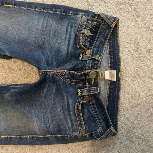 True Religion blå bootcut jeans - Snygga blå jeans från True Religion med bootcut passform och klassiska kontrastsömmar. Jeansen har detaljerade ficklock bak med tryckknappar och slitningar framtill för en cool look. Tillverkade i jeansmaterial med guldfärgade sömmar och unika knappar.skriv vid frågor!💕
