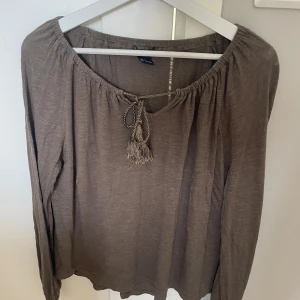 Beige blus med knyt från Lindex - Säljer en beige blus från Lindex med långa ärmar och snygg knytning med tofs framtill vid halsen. Blusen har en avslappnad passform och är tillverkad i ett mjukt, tunt material som känns skönt mot huden.