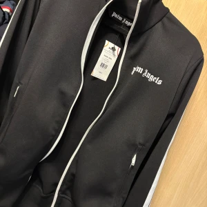 Svart zip hoodie från Palm Angels - Svart hoodie från Palm Angels med vit dragkedja och vita ränder längs ärmarna. Tröjan har hög krage, fickor med dragkedja och logotypen broderad på bröstet. Snygg streetwear-vibe och perfekt för dig som gillar stilrena detaljer.