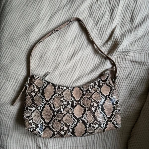 Väska i ormmönster  - Cool axelväska från NLY Accessories i beige och bruna toner med ormmönster. Väskan har justerbart axelband med spänne och stängs med dragkedja. Perfekt storlek 30x20 cm