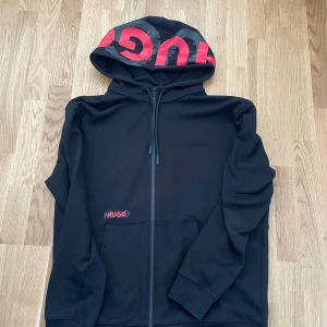 Svart hoodie från Hugo med röd logga - Svart hoodie från Hugo med stor röd logga på huvan och mindre logga på bröstet. Tröjan har dragkedja framtill, snörning i huvan och två fickor. Materialet är mjukt och känns som polyester, perfekt för chill eller streetwear. Snygg och modern design med lång ärm.