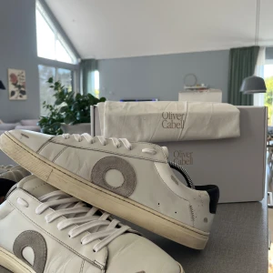 Vita sneakers från Oliver Cabell - Snygga vita sneakers från Oliver Cabell med grå mockadetalj i form av en cirkel på sidan. Skorna har klassisk låg profil, vita snören och svart hälparti. 