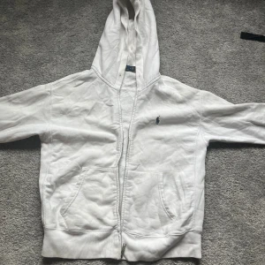 Vit hoodie från Polo Ralph Lauren - Vit hoodie från Polo Ralph Lauren med dragkedja och klassisk blå logga på bröstet. Tröjan har huva, känguruficka och ribbade muddar. Perfekt för en chill och stilren look. Tillverkad i mjuk bomull för extra komfort. Den är lite mindre i passformen, därför står det M, men passar mer som S eller XS. Den har lite färg på ena armen
