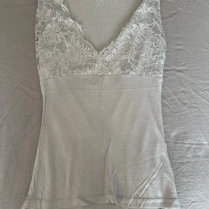 Ljusgrå spetsdetaljerad linnetopp - Supersöt ljusgrå linnetopp med breda axelband och v-ringning. Överdelen har vackra spetsdetaljer med blommönster och fransig kant, medan nederdelen är i mjukt, stretchigt tyg. Perfekt att styla med jeans eller kjol för en trendig look.