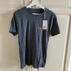Mörkgrå Hallifornia t-shirt från Dedicated - Snygg mörkgrå t-shirt från Dedicated med orange Hallifornia-tryck fram och stort handtecken-tryck bak. Klassisk rund hals och korta ärmar. Tillverkad i mjuk bomull som känns skön mot huden. Passar till tjej och kille. Storlek S. Helt ny och etiketten sitter kvar.