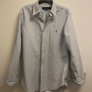 Randig skjorta från Ralph Lauren - Snygg slim fit skjorta från Ralph Lauren i vitt och blått med tunna vertikala ränder. Klassisk krage, knappar framtill och broderad logga på bröstet. Tillverkad i mjuk bomull och har lång ärm. Perfekt för dig som gillar stilrena detaljer.