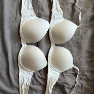 Vit bh med rosett - Säljer två vita bh:ar med vadderade kupor och liten rosett framtill. Justerbara axelband och klassisk knäppning där bak. 