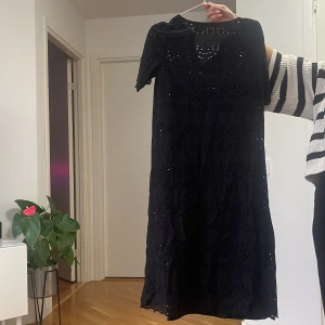 Svart långklänning med spetsdetaljer - Svart långklänning med korta ärmar och v-ringning. Klänningen har fina spetsdetaljer och broderade mönster över hela tyget som ger en cool och elegant känsla. Perfekt för dig som gillar en stilren look med det lilla extra.