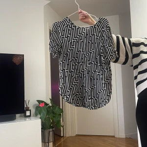 Svartvit mönstrad blus med kort ärm - Säljer en svartvit blus med grafiskt mönster och rund hals. Toppen har korta ärmar och en lös passform, perfekt för dig som gillar en stilren men ändå cool look. Passar till både jeans och kjol för en trendig outfit.
