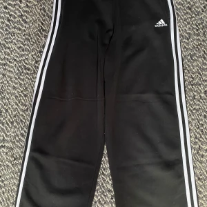 Svarta Adidas mjukisbyxor med vita ränder - Svarta mjukisbyxor från Adidas med klassiska vita ränder längs sidorna och Adidas-logga på höften. Byxorna har resår i midjan och är tillverkade i mjukt material som passar perfekt för chill eller träning. Snygg och enkel design som alltid funkar.