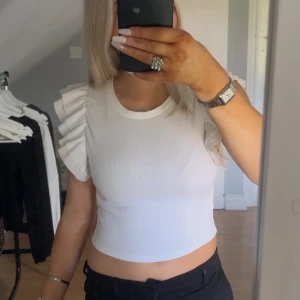 Vit ribbad topp med volangärm Zara - Superfin vit ribbad topp från Zara med korta ärmar och stora volangdetaljer på axlarna. Croppad modell som sitter snyggt och ger en trendig look. Använd ett par gånge för länge sedan men inte ofta. Inget fel