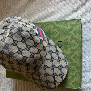 Gucci keps - Snygg keps från Gucci i beige med mörkblått GG-monogram över hela kepsen och en ikonisk röd-blå rand på sidan. Justerbar rem bak i skinn och insida i brunt tyg. Perfekt för dig som vill sticka ut med lyxig streetstyle.