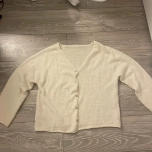 Beige stickad kofta med knappar - Mysig beige stickad kofta med v-ringning och knäppning framtill. Koftan har långa ärmar och en enkel, avslappnad passform. Perfekt att slänga på sig över en t-shirt eller linne för extra värme. Egen stickad 