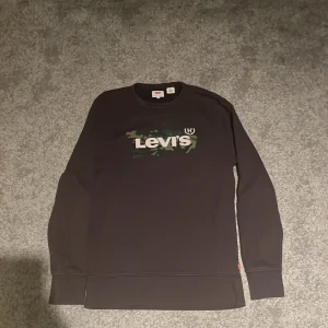 Svart Levi's sweatshirt med camo-logga - Svart sweatshirt från Levi's med klassisk rund hals och lång ärm. Framsidan har en stor Levi's-logga med camouflage-mönster i grönt och vitt. Tröjan är i mjuk bomull och har ribbade muddar vid ärmslut och nederkant. Perfekt för en chill och stilren look.