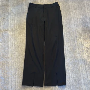 Svarta kostymbyxor med slits - Stilrena svarta kostymbyxor med raka ben och snygga slitsar framtill vid bensluten. Byxorna har hällor för bälte och sidfickor. Perfekta för en clean och trendig look. Materialet känns mjukt och följsamt.