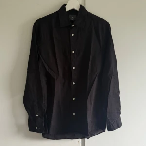 Svart linneskjorta från H&M - Svart skjorta från H&M i regular fit med klassisk krage och vita knappar. Tillverkad i en skön mix av 52% linne och 48% bomull, perfekt för en clean och stilren look. Långärmad modell som funkar året runt. Använd en gång