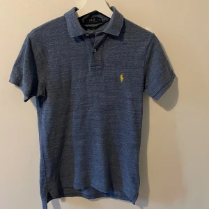 Blå Polo Ralph Lauren t-shirt slim fit - Snygg blå t-shirt från Polo Ralph Lauren i slim fit-modell. Klassisk pikékrage med två knappar och korta ärmar. Ikonisk gul broderad logga på bröstet. Mjuk bomullskvalitet som känns skön mot huden. Perfekt för dig som gillar stilren och sportig look. Priset kan diskuteras