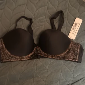 Svart spetsigt bh  från Fyeya - Snygg svart bh från Freya med vadderade kupor och avtagbara axelband. Sidorna är dekorerade med svart spets över beige tyg, vilket ger en lyxig känsla. Perfekt för att lyfta vilken outfit som helst med en touch av elegans.