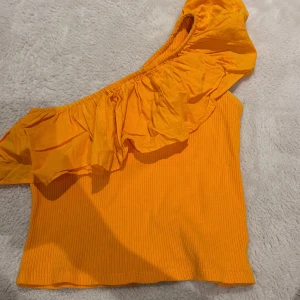 Orange one shoulder topp från Zara - Säljer en superfin orange one shoulder topp från Zara. Toppen är ribbad och har en bred volang längs den asymmetriska axeln. Perfekt för dig som vill sticka ut med en färgglad och trendig look. Materialet är mjukt och stretchigt för en bekväm passform.