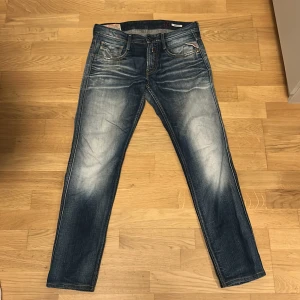 Replay Anbass jeans - Snygga Replay Anbass jeans i blå tvätt med slitningar och ljusa partier. Klassisk femficksmodell med raka ben och normal passform. Tuffa detaljer på bakfickan och tydliga kontrastsömmar. Perfekta för dig som gillar en avslappnad men trendig look.