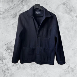 Svart overshirt från A Day's March XS - Svart overshirt från A Day's March i storlek XS. Jackan har tre stora fickor framtill, klassisk krage och knäppning med knappar. Materialet är mjukt och känns som ull, perfekt för lager-på-lager. Enkel och stilren design som funkar till många outfits.